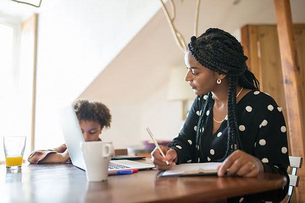 A woman writing notes at a table while a child uses a laptop beside her